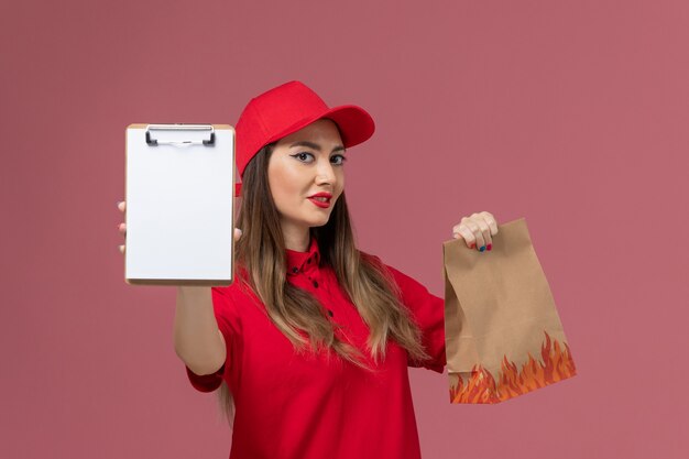 Correio feminino de uniforme vermelho segurando o bloco de notas e o pacote de comida no chão rosa. Empresa de entrega de serviço