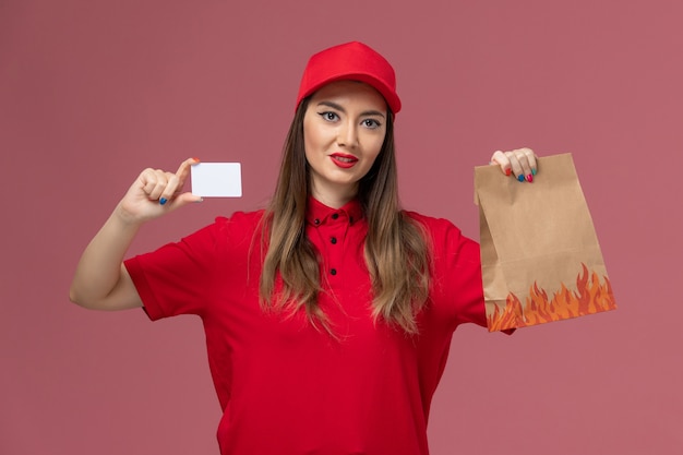 Correio feminino de uniforme vermelho segurando um cartão branco e um pacote de comida no fundo rosa.