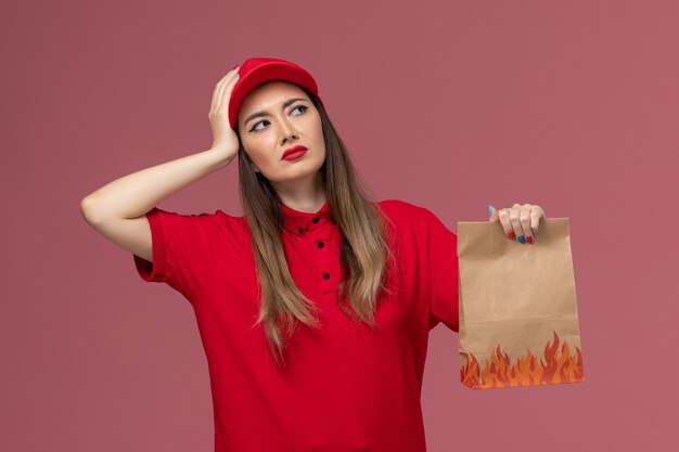 Correio feminino de uniforme vermelho segurando um pacote de comida de papel tocando sua cabeça no fundo rosa.