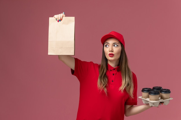 Correio feminino de uniforme vermelho segurando xícaras de café com pacote de comida em fundo rosa claro uniforme de trabalho de entrega de serviço