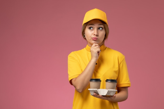 Correio feminino de vista frontal com capa amarela uniforme amarela segurando café com expressão pensativa no fundo rosa uniforme trabalho de entrega