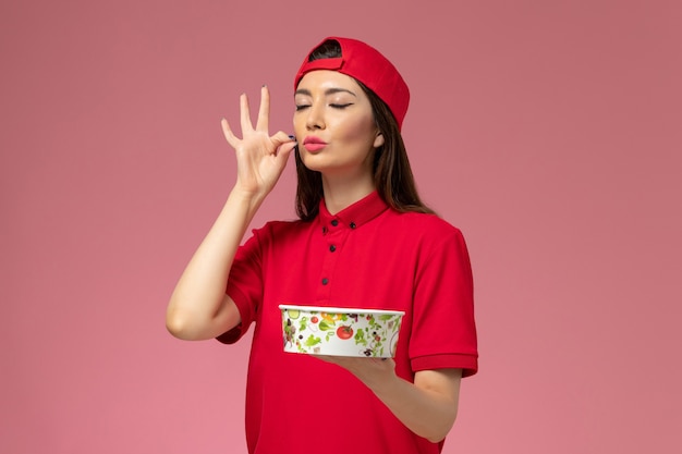 Correio feminino de vista frontal com capa de uniforme vermelho com tigela de entrega nas mãos na parede rosa claro, funcionário de entrega de uniforme