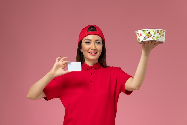 Correio feminino de vista frontal com capa uniforme vermelha com tigela de entrega e cartão branco nas mãos na parede rosa claro, trabalho de empregado de entrega de uniforme