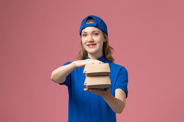 Correio feminino de vista frontal com uniforme azul e capa segurando pequenos pacotes de comida de entrega na parede rosa, empresa de serviço de uniforme de trabalho de entrega