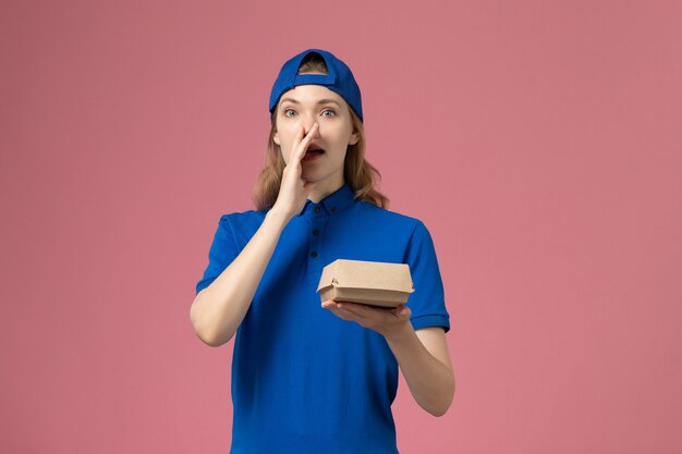 Correio feminino de vista frontal com uniforme azul e capa segurando um pequeno pacote de entrega de comida gritando na parede rosa, empresa de serviço de uniforme de entrega