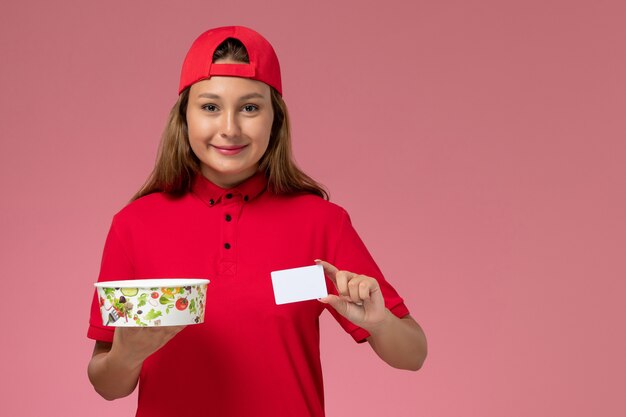 Correio feminino de vista frontal com uniforme vermelho e capa segurando a tigela de entrega e o cartão na parede rosa, serviço de entrega de uniforme