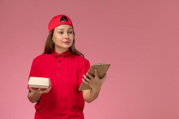 Correio feminino de vista frontal com uniforme vermelho e capa segurando um pequeno pacote de comida de entrega e um bloco de notas na parede rosa, empresa de serviço de entrega de uniforme de trabalho