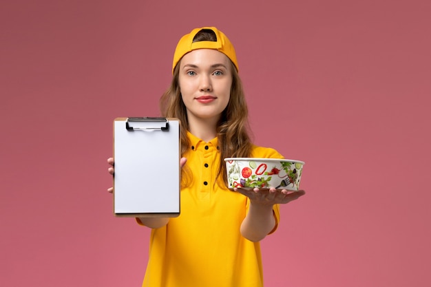 Foto grátis correio feminino de vista frontal em uniforme amarelo e capa segurando a tigela de entrega e o bloco de notas na parede rosa claro serviço de entrega de uniforme da empresa trabalho