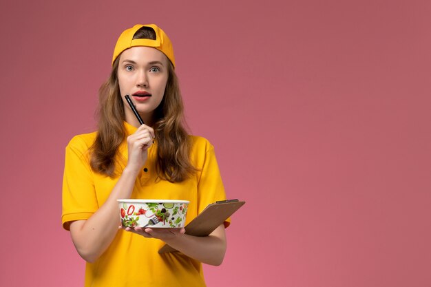 Correio feminino de vista frontal em uniforme amarelo e capa segurando a tigela de entrega e o bloco de notas pensando na parede rosa claro serviço trabalhador entrega uniforme empresa de trabalho