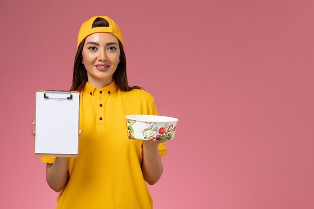 Foto grátis correio feminino de vista frontal em uniforme amarelo e capa segurando a tigela de entrega redonda com bloco de notas na parede rosa serviço uniforme entrega trabalho