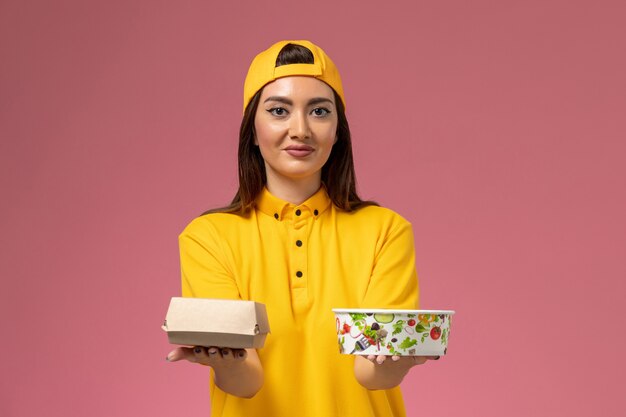 Correio feminino de vista frontal em uniforme amarelo e capa segurando o pacote de comida com tigela na parede rosa claro.