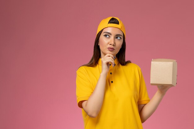 Correio feminino de vista frontal em uniforme amarelo e capa segurando pequeno pacote de comida de entrega pensando em empresa de entrega de serviço de uniforme de parede rosa claro