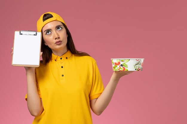 Foto grátis correio feminino de vista frontal em uniforme amarelo e capa segurando tigela de entrega redonda com bloco de notas na parede rosa serviço de entrega uniforme de trabalho