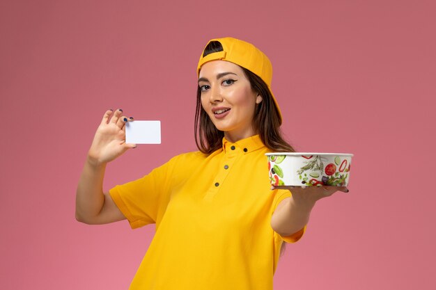 Correio feminino de vista frontal em uniforme amarelo e capa segurando tigela de entrega redonda e cartão branco na parede rosa.