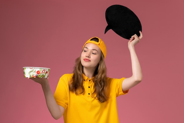 Correio feminino de vista frontal em uniforme amarelo e capa segurando uma placa preta com tigela na parede rosa empresa de entrega de uniforme de serviço