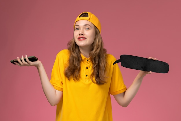 Correio feminino de vista frontal em uniforme amarelo e capa segurando uma placa preta e smartphone na parede rosa uniforme.