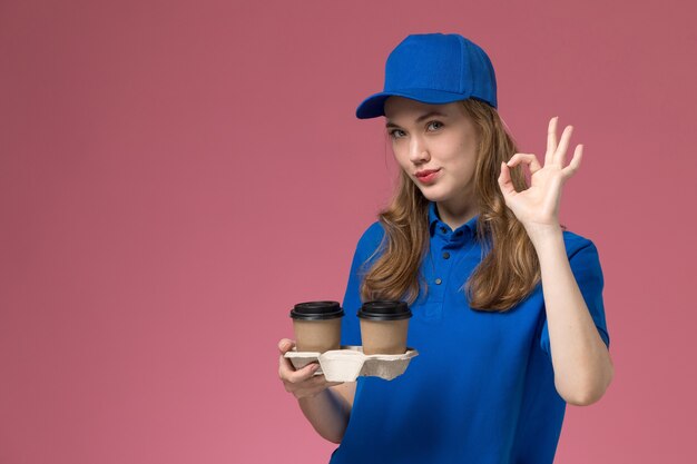 Correio feminino de vista frontal em uniforme azul segurando xícaras de café marrons, mostrando um bom sorriso no uniforme de serviço de fundo rosa, entregando trabalho da empresa