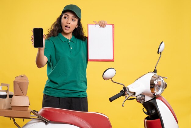 Correio feminino de vista frontal em uniforme verde com telefone e nota de arquivo no fundo amarelo.