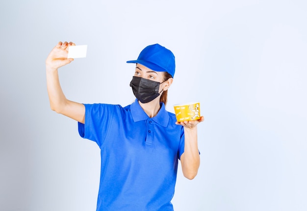 Correio feminino do restaurante de uniforme azul e máscara facial, entregando uma comida para viagem e apresentando seu cartão de visita.