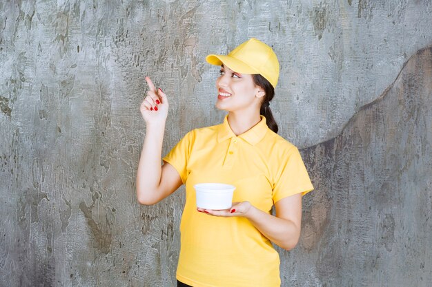 Correio feminino em uniforme amarelo, segurando uma xícara para viagem e apontando para alguém.