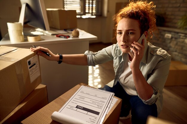 Correio feminino preocupado se comunicando por telefone celular enquanto trabalha com pacotes de entrega no escritório