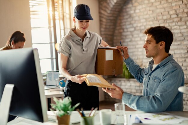 Correio feminino sorridente entregando pacote para um empresário enquanto ele está trabalhando no escritório