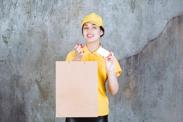 Correio feminino vestindo uniforme amarelo, entregando uma sacola de compras de papelão e apresentando seu cartão de visita.