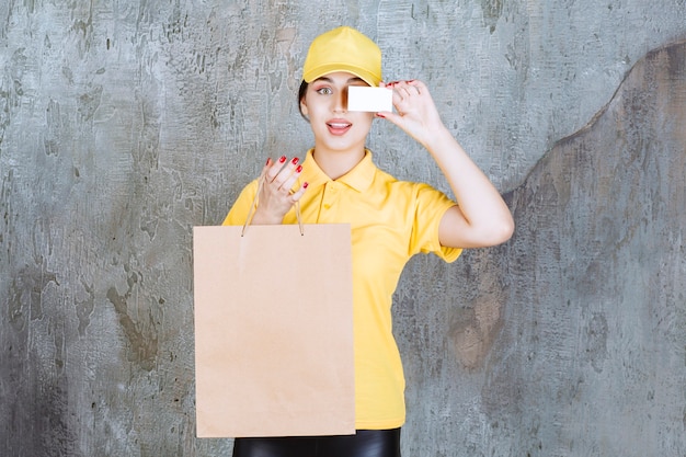 Correio feminino vestindo uniforme amarelo, entregando uma sacola de compras de papelão e apresentando seu cartão de visita.