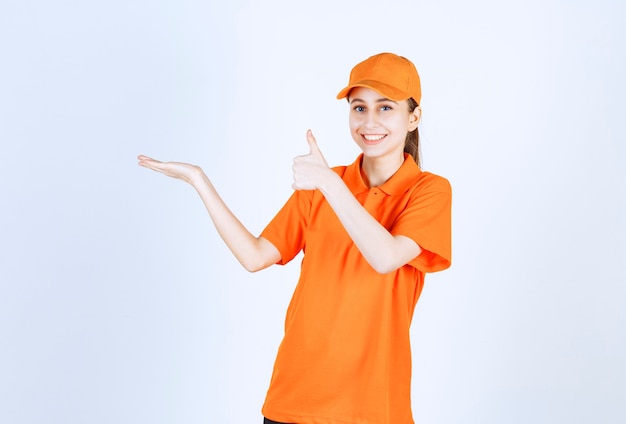 Correio feminino vestindo uniforme laranja e boné aparecendo o polegar.