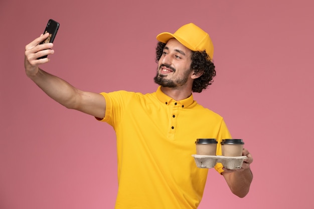 Correio masculino de uniforme amarelo segurando xícaras de café marrons tirando foto na parede rosa