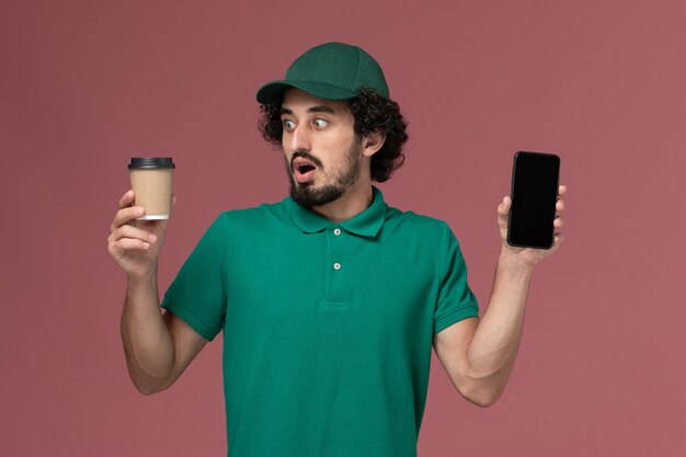 Correio masculino de vista frontal com uniforme verde e capa segurando a xícara de café de entrega com telefone no serviço de entrega uniforme de fundo rosa