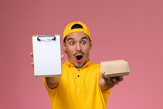 Correio masculino de vista frontal de uniforme amarelo, segurando o bloco de notas e pouco pacote de comida no fundo rosa claro.