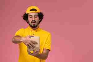 Foto grátis correio masculino de vista frontal em uniforme amarelo e capa com pouco pacote de comida de entrega nas mãos abrindo-o no fundo rosa.