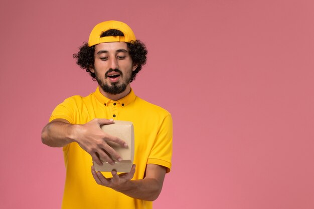 Correio masculino de vista frontal em uniforme amarelo e capa com pouco pacote de comida de entrega nas mãos abrindo-o no fundo rosa.