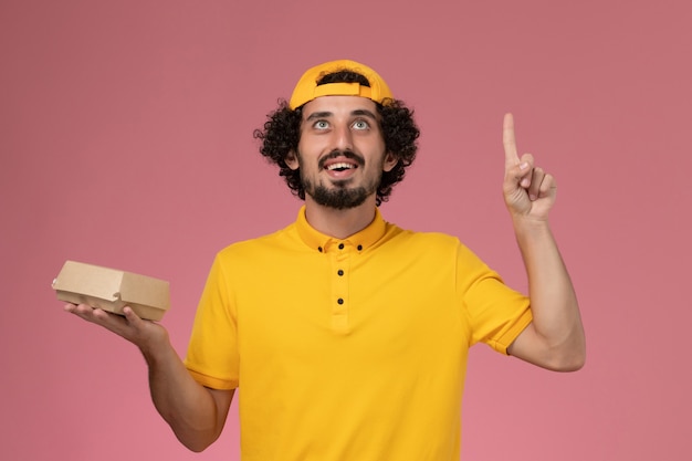 Correio masculino de vista frontal em uniforme amarelo e capa com pouco pacote de comida de entrega nas mãos sobre o fundo rosa.