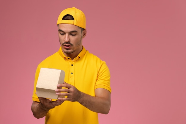 Foto grátis correio masculino de vista frontal em uniforme amarelo e capa segurando e abrindo pequenos pacotes de comida de entrega no fundo rosa.
