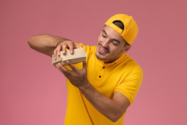 Correio masculino de vista frontal em uniforme amarelo e capa segurando o pequeno pacote de comida de entrega na mesa rosa.
