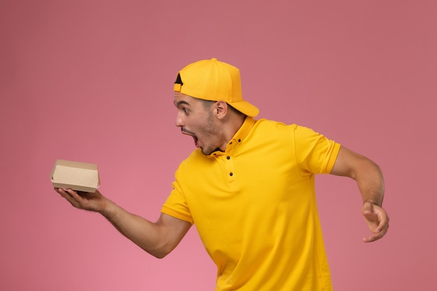 Correio masculino de vista frontal em uniforme amarelo e capa segurando pequeno pacote de comida de entrega com medo no fundo rosa claro.