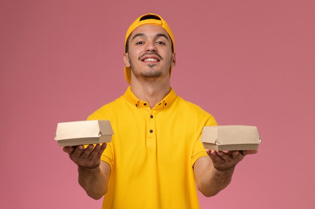 Correio masculino de vista frontal em uniforme amarelo e capa segurando pequenos pacotes de comida de entrega na mesa rosa.