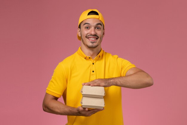 Correio masculino de vista frontal em uniforme amarelo e capa segurando pequenos pacotes de comida de entrega no fundo rosa claro.