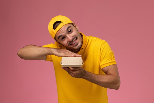 Correio masculino de vista frontal em uniforme amarelo e capa segurando um pequeno pacote de comida de entrega em fundo rosa claro.