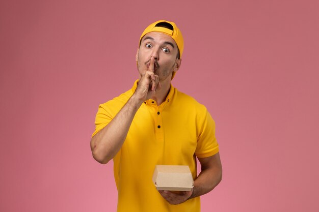 Correio masculino de vista frontal em uniforme amarelo e capa segurando um pequeno pacote de comida de entrega em fundo rosa.