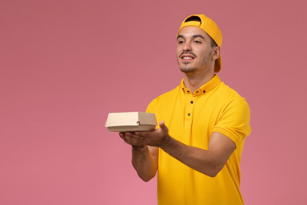 Correio masculino de vista frontal em uniforme amarelo e capa segurando um pequeno pacote de comida de entrega em fundo rosa.