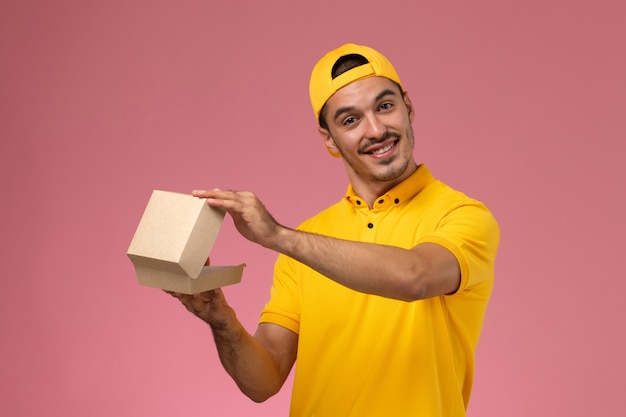 Correio masculino de vista frontal em uniforme amarelo e capa segurando um pequeno pacote de comida de entrega, sorrindo na mesa rosa.