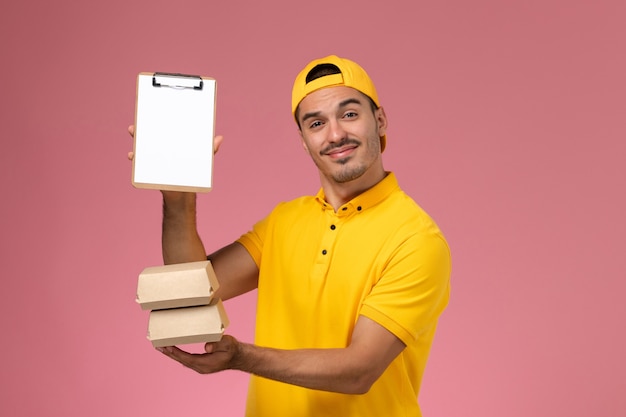 Foto grátis correio masculino de vista frontal em uniforme amarelo, segurando pequenos pacotes de comida e o bloco de notas sobre o fundo rosa claro.