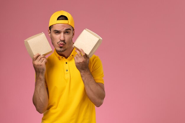 Correio masculino de vista frontal em uniforme amarelo, segurando pequenos pacotes de comida no fundo rosa.