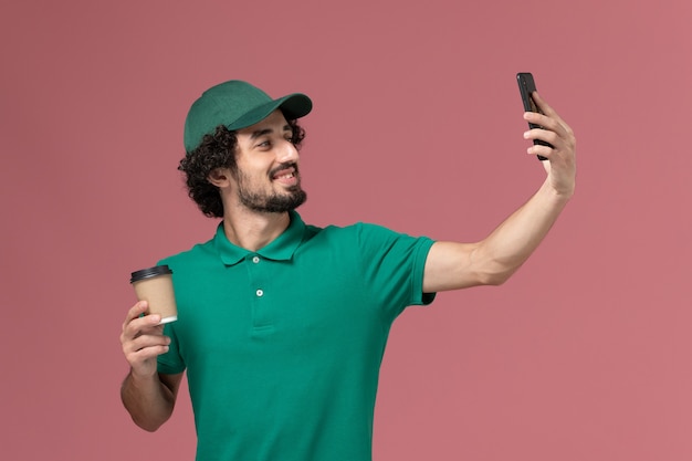 Correio masculino de vista frontal em uniforme verde e capa segurando xícara de café tomando selfie em serviço de entrega uniforme de fundo rosa