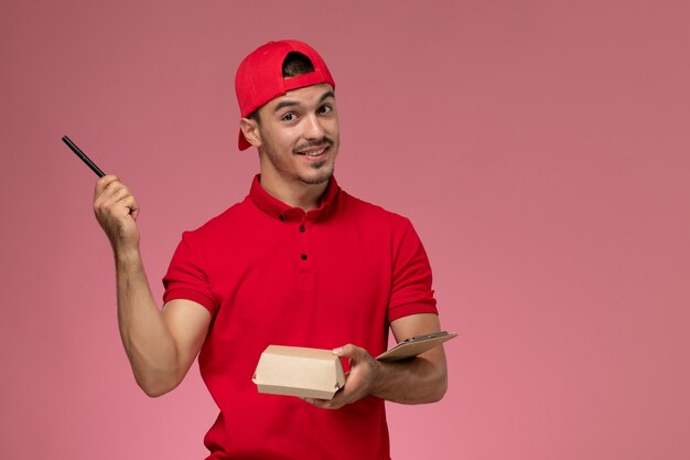 Correio masculino de vista frontal em uniforme vermelho e capa segurando o pequeno pacote de entrega com o bloco de notas escrevendo nota sobre o fundo rosa.