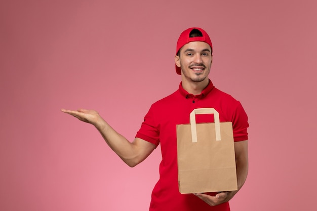 Correio masculino jovem de vista frontal na capa uniforme vermelha, segurando o pacote de comida de papel no fundo rosa claro.