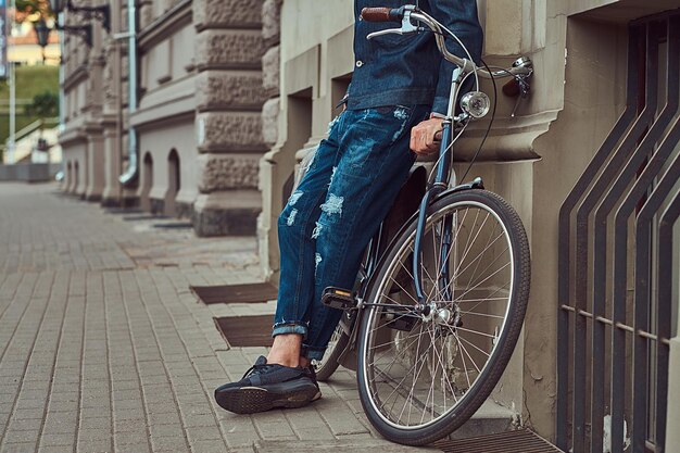 Cortada a imagem de um homem elegante em roupas elegantes, encostado a uma parede com bicicleta da cidade em uma rua.